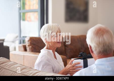 Heim, Liebe und altes Paar auf Sofa, Geschenk und Bindung zusammen mit Jubiläum. Wohnung, Rentner und Seniorin auf Couch, Mann und Geschenk mit Stockfoto