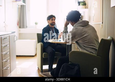Rückansicht eines lockhaarigen Teenagerschülers, der sich auf den Ellenbogen gestützt hat, während er mit dem Berater im Oberschulamt sitzt Stockfoto