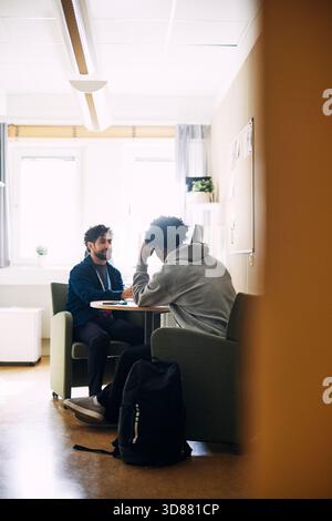 Rückansicht eines Teenager-Schülers, der sich auf den Ellenbogen gestützt hat, während er mit dem Betreuer im Oberschulamt sitzt Stockfoto
