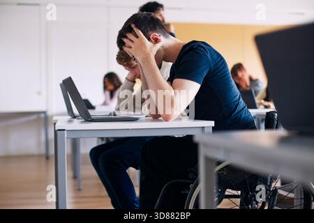 Gestresster männlicher High School Schüler sitzt im Rollstuhl mit Kopf in den Händen im Klassenzimmer Stockfoto