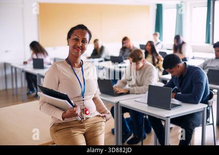 Porträt einer selbstbewussten Bildungsexpertin, die Bücher hält, die am Tischrand im Klassenzimmer der High School sitzen Stockfoto