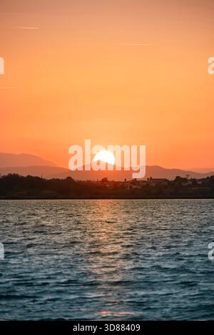 Vertikaler Blick auf den Sonnenuntergang oder -Aufstieg über Hügel und Küstengebäude mit dramatischem orangefarbenem Licht, das auf dem Meer reflektiert Stockfoto