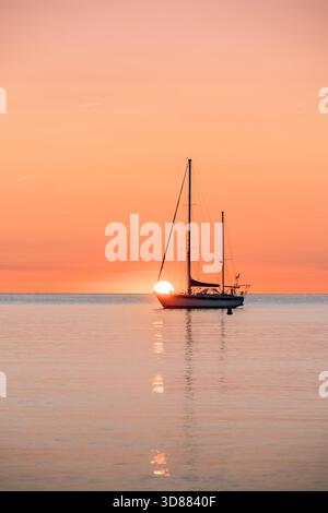 Vertikales Porträt eines Segelbootes, das auf einem ruhigen Meer vor Anker liegt, mit einer Silhouette vor dem hellen orange-pastellfarbenen Himmel während Sonnenauf- oder -Untergang Stockfoto