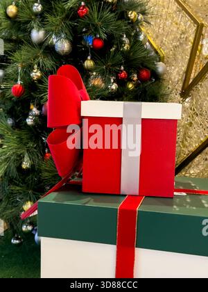 Drei gestapelte Geschenkboxen in Rot, Grün und weiß, verziert mit Bändern und einer Schleife, werden vor einem Weihnachtsbaum platziert. Stockfoto