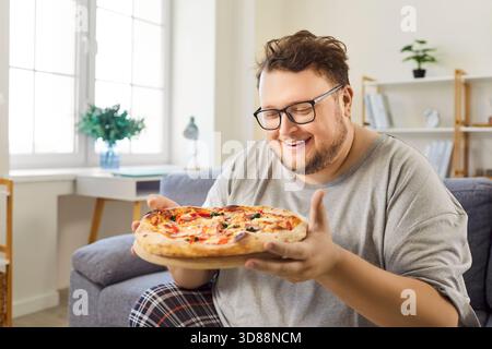 Übergewichtiger Mann, der Pizza mit fröhlichem Ausdruck und Lächeln hält, auf dem Sofa sitzt Stockfoto