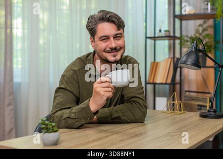Ein Mann mittleren Alters hält zu Hause eine Tasse Kaffee und genießt einen entspannenden Moment und fühlt Ruhe. Junger Erwachsener am Tisch atmet langsam eine ruhige Stimmung Zufriedenheit und ruht sich aus, bevor er heute seine Aufgaben fortsetzt Stockfoto