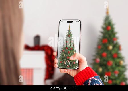 Frau fotografiert einen geschmückten Weihnachtsbaum mit Smartphone, hält festliche Ornamente, warme Feiertagsatmosphäre und gemütliche Winterszene fest Stockfoto