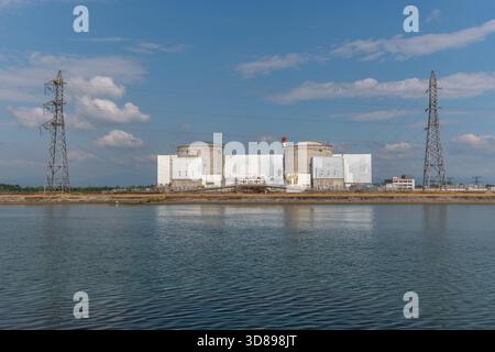 Eine große industrielle Atomanlage liegt an einer ruhigen Uferpromenade. Zwei hohe zylindrische Strukturen dominieren die Skyline, wobei Stromleitungen über dem Himmel verlaufen und entfernte Wolken den blauen Himmel verstärken. Fessenheim, Oberrhein, Elsass, Frankreich, Europa. Stockfoto
