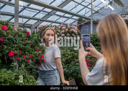 Die junge Frau steht inmitten farbenfroher Blumen in einem Gewächshaus, während ihre Tochter im Teenageralter ein Foto macht, das ihre fröhliche Beziehung und ihre gemeinsame Ex hervorhebt Stockfoto