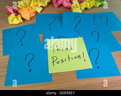 Denken Sie Positiv! Bei Fragen – Grün „Positiv Denken!“ Haftnotiz, umgeben von blauen Fragezeichen und zerknittertem Papier, symbolisiert das Problem... Stockfoto