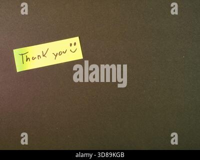 Dankeschön auf dunklem Hintergrund - gelbe Haftnotiz mit einer handgeschriebenen "Danke :)"-Nachricht auf einem strukturierten dunkelgrünlich-braunen Hintergrund, Symbolen Stockfoto