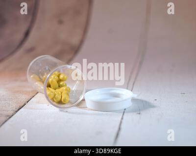 Gelbe Kapseln aus Pillenflasche verschütten - gelbe ovale Kapseln verschütten sich aus einer transparenten Arzneimittelflasche aus Kunststoff neben der weißen Kappe auf einer sauberen, lila Stockfoto