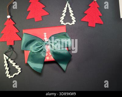 Festliche Geschenkbox und Weihnachtsdekoration – Rote Geschenkbox mit grüner Schleife und Band, umgeben von festlichen Weihnachtsbaumschmuck auf dunklem Hintergrund. Leistung Stockfoto