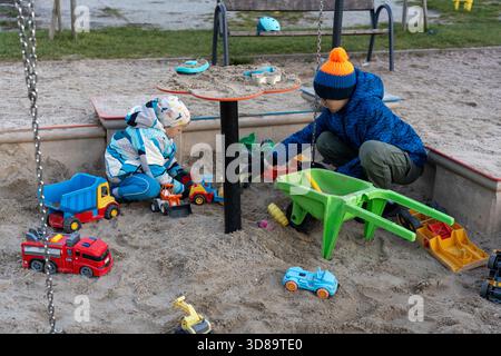 Zwei Brüder spielen am Herbsttag mit Spielzeugwagen und Schubkarre im Sandkasten auf dem Spielplatz. Vorstellung von Geschwistern fantasievolles Spiel, Outdoor-Kindheit f Stockfoto