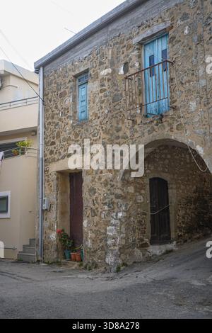 Vertikale Aufnahme eines historischen Steinhauses in Melampes, Kreta, mit einem bogenförmigen Eingang, rostigem Balkongeländer und verblassten blauen Fensterläden Stockfoto