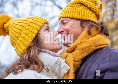 Ein fröhliches Paar mittleren Alters in gelben Hüten und Tüchern genießt einen kurzen Moment in einer herbstlichen Umgebung, umgeben von lebhaften Herbstblättern und einem warmen Atmo Stockfoto
