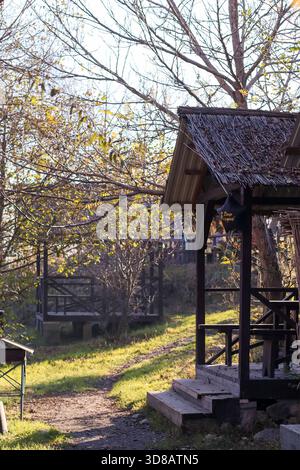 Rustikale Holzpavillons mit Strohdächern entlang eines Kiesweges in einem üppig grünen Park bei Sonnenschein im Herbst. Stockfoto