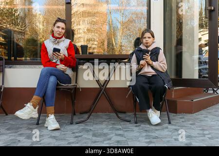 Frau in rotem Pullover hält Smartphone, sitzt neben einem Mädchen, das aus der Teenagerin trinkt, am Kaffeetisch im Freien in urbaner Umgebung. Stockfoto