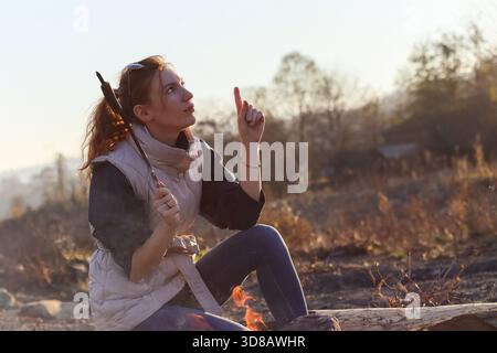 Weibchen im mehrschichtigen Outfit Gesten mit erhobenem Finger, Wurst auf Stock in der Nähe von Flammen in landschaftlich schöner Herbstnähe von fröhlicher junger Frau mit Pferdeschwanz Stockfoto