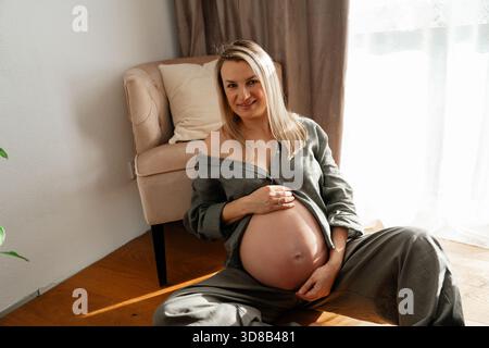 34 Wochen, Ein sanfter Mutterschaftsmoment mit einer schwangeren Frau, die bei hellem Tageslicht auf dem Boden in der Nähe eines Sofas sitzt. Ruhige Töne und eine entspannte Haltung sorgen für einen Stockfoto