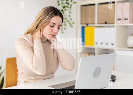 Müde Frau, die den Hals berührt und Muskelschmerzen am Schreibtisch spürt. Stockfoto