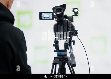 Filmeinrichtung mit einer professionellen Videokamera auf einem Stativ, das vor eine helle Kulisse gerichtet ist, während ein Bediener in der Nähe in einer Innenproduktion steht Stockfoto