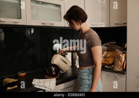 Eine Frau gießt heißes Wasser aus einem Wasserkocher in eine Teekanne aus Glas und bereitet in einer ruhigen Küche ein warmes Getränk zu. Stockfoto