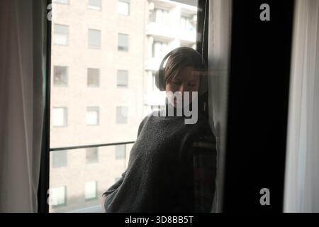 Eine Frau mit Kopfhörern lehnt sich mit sanfter Reflexion an ein Fenster und schafft eine nachdenkliche und introspektive Szene. Stockfoto