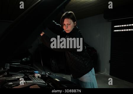 Eine Frau öffnet die Motorhaube und überprüft den Motorraum in einer unterirdischen Garage. Der Moment zeigt die routinemäßige Fahrzeuginspektion und das verantwortliche Fahrzeug Stockfoto