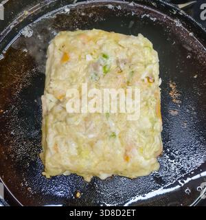 Goldbraun gebräuntes Okonomiyaki kocht auf einer Kochplatte und zeigt seine reichhaltige herzhafte Textur, Kohl-Mischung und traditionelle japanische Street Food-Stil. Stockfoto
