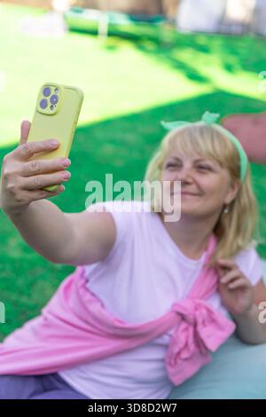 Eine blonde Frau in lässiger, heller Kleidung sitzt auf einem Rasen, nimmt ein Selfie auf oder plaudert im Freien durch Videoanrufe Stockfoto