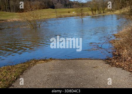Eine Landstraße endet abrupt in einem überfluteten Gebiet, umgeben von einer ruhigen natürlichen Landschaft während eines Hochwasserereignisses, das durch saisonale Veränderungen verursacht wird. Stockfoto