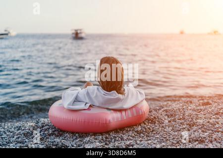 Strand, Sonnenuntergang, Urlaub. Person, die am Kieselstrand im Donut Float sitzt, auf das ruhige Meer blickt, friedlicher Sommerabend. Stockfoto