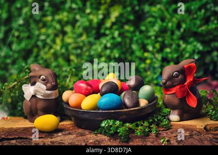 Schokolade Ostereier mit Hase und Ei Bonbons auf einem Holztisch im Hof mit grünen Frühling Pflanzen in Der Hintergrund Stockfoto