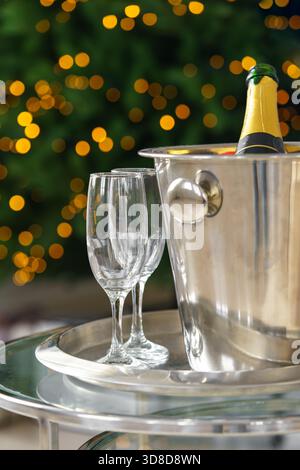 Champagnerflasche in Metall Eiskübel mit zwei leeren Gläsern auf Tablett auf leuchtenden Weihnachtsbaumlichtern Hintergrund. Festliche Winterurlaubsatmosphäre pro Stockfoto