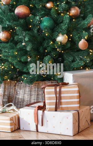 Schöne Weihnachtspapiertüten und Geschenkboxen, die in elegantes Papier eingewickelt sind, unter grünem Weihnachtsbaum mit warmen Lichtern. Gemütliches Winterurlaubsambiente Stockfoto