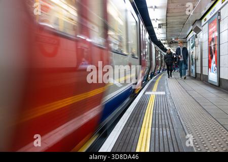 Pendler gehen neben dem Moving Tube Train im Londoner U-Bahn-System. London, Vereinigtes Königreich, 10. Dezember 2023 Stockfoto