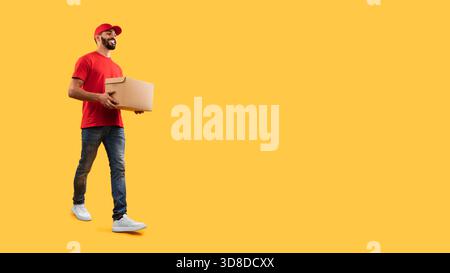 Lächelnde Zustellperson, die mit einem Paket vor einem leuchtend gelben Hintergrund läuft Stockfoto