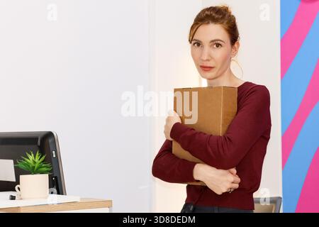 Junge Frau, die Papppaket in einem modernen Raum mit Schreibtischanlage und Monitor umschließt Stockfoto