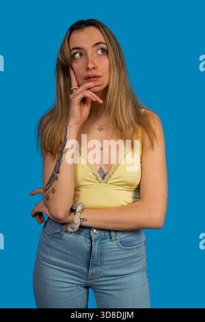 Eine junge Frau mit Tattoos, die aufmerksam denkt und auf blauem Hintergrund aufblickt Stockfoto