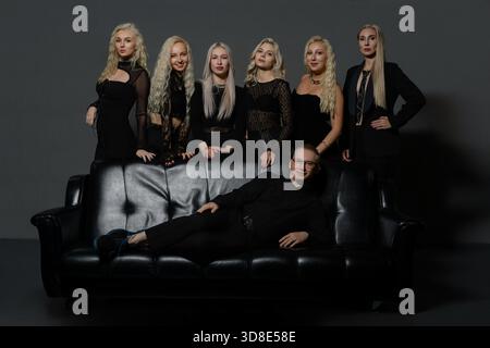 Sieben Personen mit langen, hellen Haaren tragen schwarze Kleidung. Sie sind auf einer schwarzen Couch in einem Studio-Ambiente angeordnet und zeigen verschiedene stilvolle Posen und Ausdrucksformen. || Modell freigegeben Stockfoto