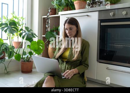Eine interessierte Freiberuflerin sitzt auf dem Boden und arbeitet zu Hause mit Pflanzen auf den Knien am Laptop. Stockfoto