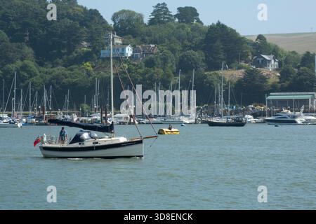 Kleines Segelboot auf ruhigem Wasser in einem belebten Hafen mit verankerten Yachten und Hügelgebäuden im Hintergrund, die die Freizeit an der Küste und die Segel veranschaulichen Stockfoto