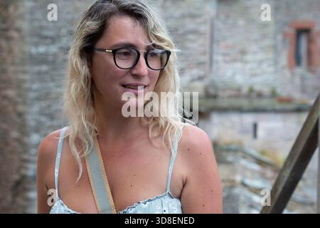 Porträt einer Frau mit Brille, die draußen vor einer verwitterten Steinmauer steht, offenes natürliches Licht, geringe Schärfentiefe. Stockfoto