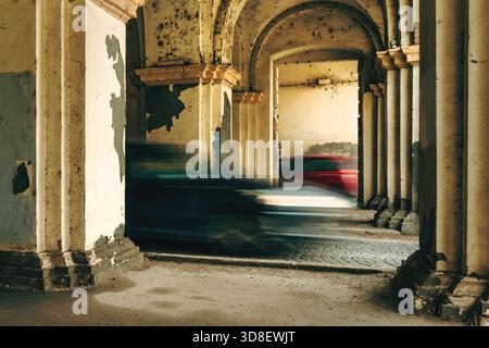 Bewegungsunscharfe Fahrzeuge, die durch eine verwitterte, bogenförmige Passage rasen und den modernen Verkehr mit veralteter, zerbröckelnder Architektur kontrastieren. Selektiver Fokus. Stockfoto