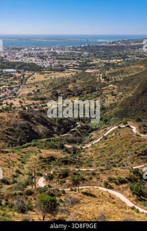 Vertikaler Hochwinkelblick vom Naturpark Arrábida, Portugal. Eine gewundene Straße schlängelt sich durch trockene Hügel, mit der Stadt Setúbal und der Küste. Stockfoto