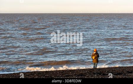Frau, die am Strand steht und den Ozean und die fernen Windräder bei Sonnenuntergang betrachtet Stockfoto