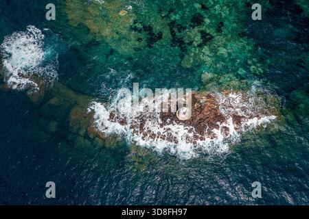 Atemberaubende Drohnenaufnahme einer zerklüfteten felsigen Küste mit türkisfarbenem Wasser und weißer Brandung, mit einem kleinen Leuchtturm an einem verwitterten Steg, Sardinien, Italien Stockfoto