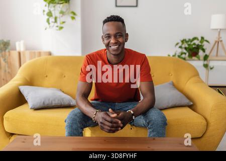 Fröhlicher afrikanischer Mann, der in die Kamera lächelt, während er zu Hause bequem auf dem Sofa sitzt Stockfoto
