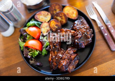 Schweinerippchen in dunkler Kruste gebraten und Beilage mit gebratenen Kartoffeln und einer Handvoll frischem Gemüse Stockfoto